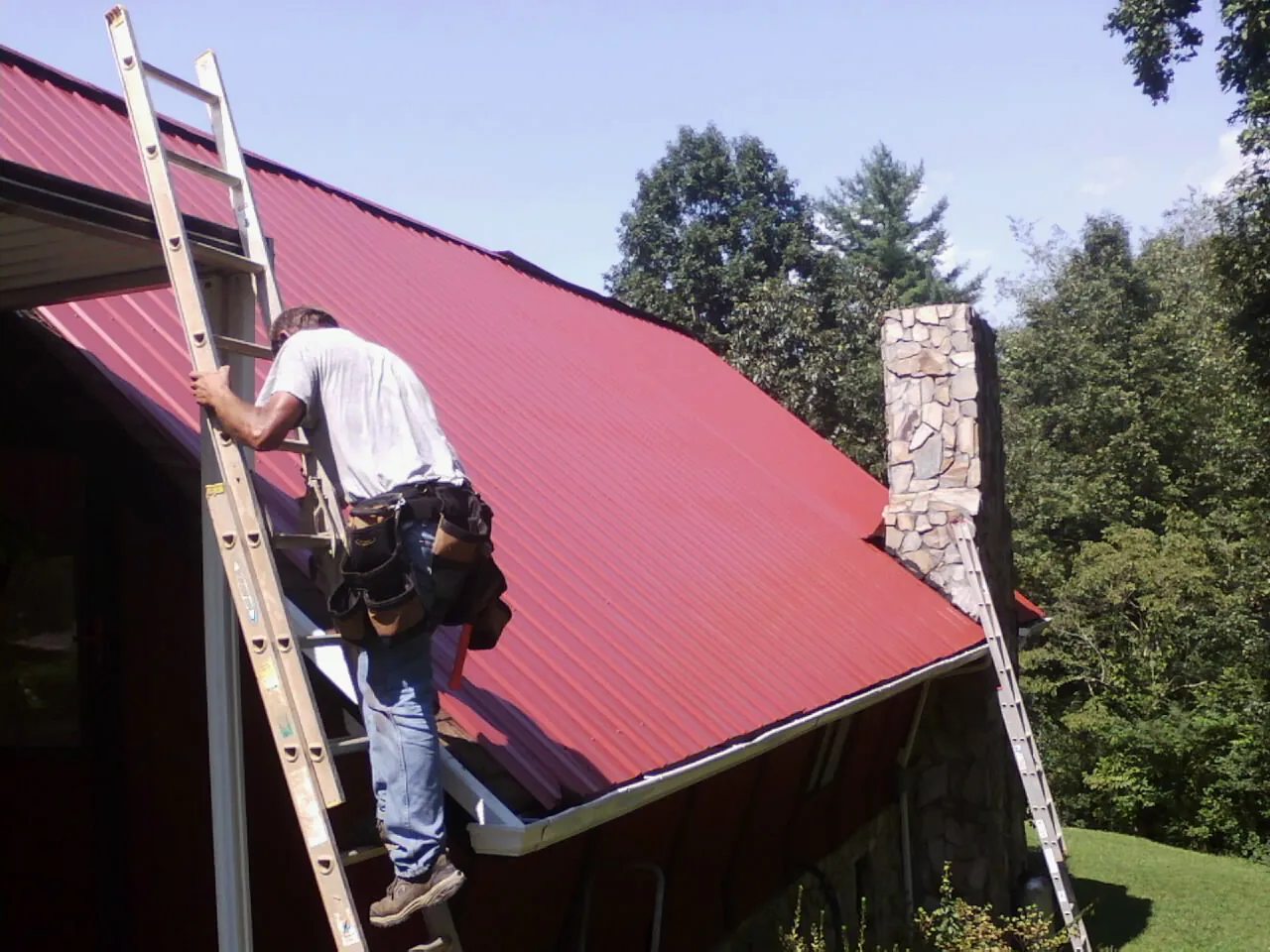 Roofer on a ladder beside a red metal roof and stone chimney — Soffit & Fascia Repair by Roofing Brothers in French Gulch